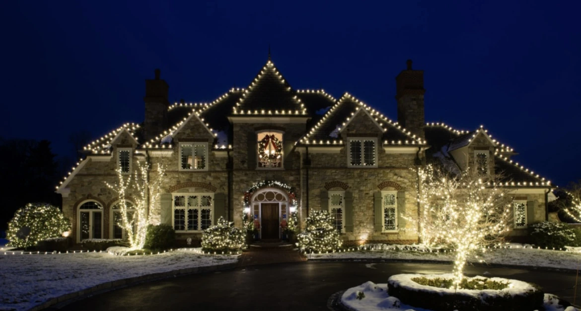 A grand Corpus Christi home outlined in warm white Christmas lights at night with wrapped trees and a snowy drive